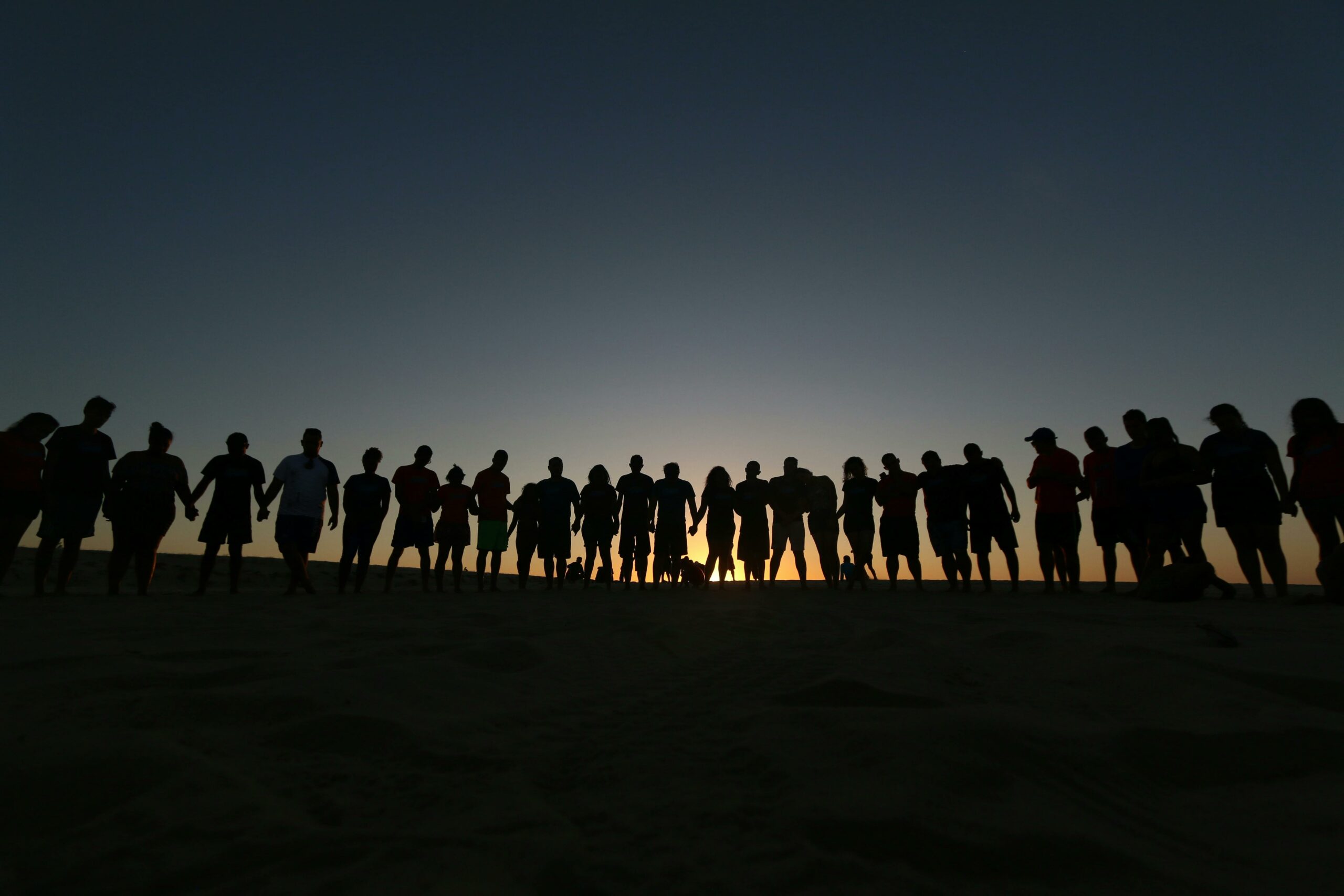 A large group holding hands as silhouettes during sunset, symbolizing unity and friendship.